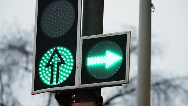 Close-up of LED traffic lights showing green straight-ahead and right-turn arrows on a pole at a city intersection, signaling allowed directions for vehicles under overcast daytime conditions