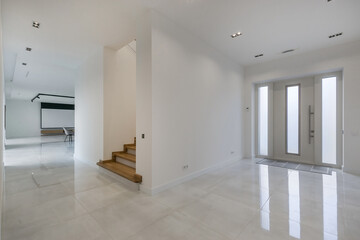 A spacious, bright hall with an entrance door and a staircase with wooden steps.  White floor tiles. Unfurnished.