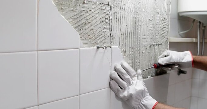 Construction worker demolishing and removing ceramic tiles in bathroom wearing protective gloves, close up