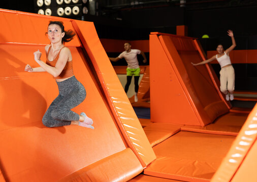 Happy emotional woman in fitness clothes jumping, pushing off from angled surface in bustling indoor trampoline park
