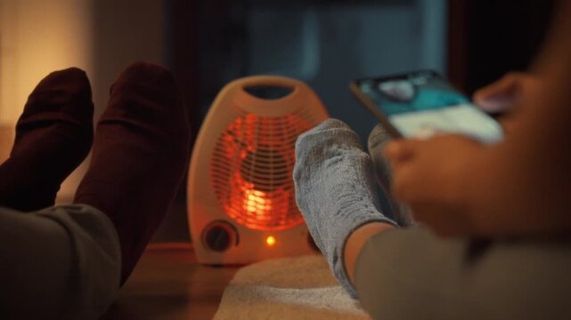 A young couple sits cozily beside a portable heater, warming their sock-clad feet on a chilly evening. The woman holds a phone, enjoying the comfort and silence of their heated house