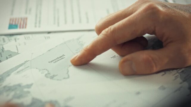 An official's emphatic gesture highlights Greenland on a large world map, surrounded by documents discussing the arctic island's sovereignty and autonomy in relation to Denmark