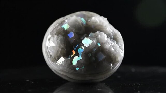 Rotating polished geode crystal with iridescent minerals and reflective surface on dark background