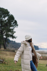Winter outdoor scene with a person in a puffy coat walking through a grassy field, wearing a beanie and casual attire under a cloudy sky.
