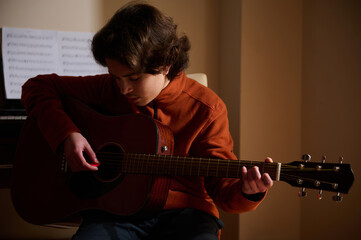 Obraz premium Teenage Boy Playing Acoustic Guitar In Warm Room, Focused On Music Hobby