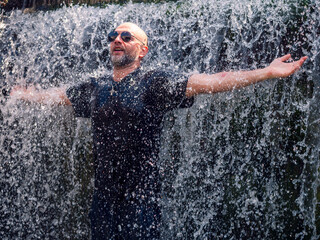 Fototapeta premium A man is standing in a waterfall, with his arms outstretched. Enjoy cold water on hot summer day. Male model in black shirt in his 40s or 50s with slim athletic body type. Have fun and cool down.
