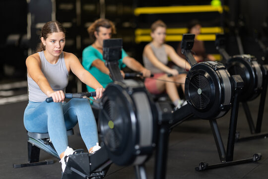 Young woman with sports leggings and a top does a cardio workout on a rowing machine. Crossfit group training participants perform a circuit training session for endurance.
