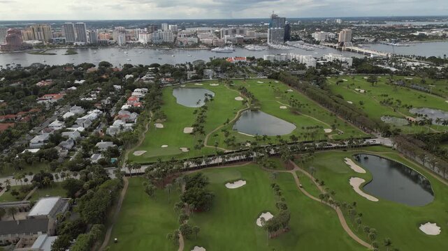 Aerial View of Luxury Tropical Golf Course with Emerald Fairways, Water Hazards, and Coastal City Skyline Backdrop featuring Waterfront Marina and Bridge.