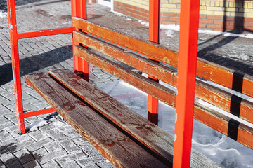 Empty red bus stop bench in winter city street. Wooden seat with metal frame covered with frost and...