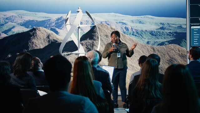 Black female speaker does wind power presentation at business forum with wind turbine machine and rotation. Addressing listeners during public speaking event focused on alternative energy.
