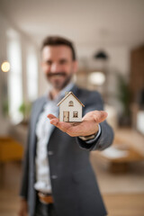 Real estate business market, house home building purchase sale. A man in a suit holding a miniature house in his hand, with a blurred background suggesting a real estate setting.