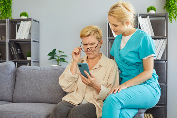 Senior woman using mobile phone with help of caregiver at home. Elderly lady with poor eyesight...
