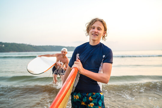 Portrait of a teen boy with a surfboard after surfing with his father. They are smiling and walking out of the water. Family active vacation concept.