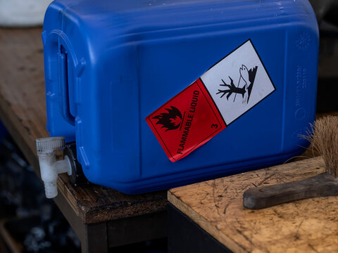 Blue plastic container with flammable liquid and environmental hazard warning labels on wooden workbench