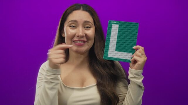 Woman smiling broadly points finger at green learner sign while holding it in purple studio; learning driving confidence growth.