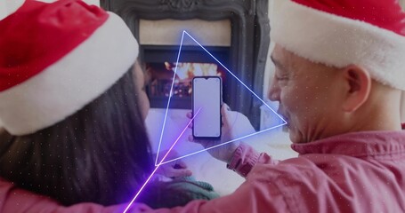 Naklejka premium Sitting couple wearing Santa hats holding smartphone showing blank screen in living room, fireplace