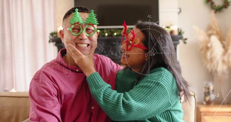 Naklejka premium Smiling couple cupping chin, wearing green knit sweater and red shirt with novelty glasses at home