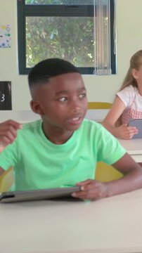 Vertical video: Raising hand boy in green tee answering teacher prompt in classroom, holding tablet