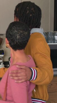 Vertical video: Gesturing man in yellow jacket guiding woman in pink top to take pink cup at cafe