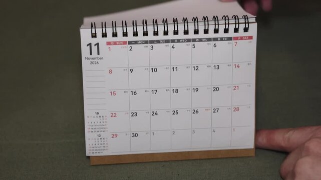 Wide close-up of a desk calendar page changing from October 2026 to November 2026. Hand flips the paper forward while the camera remains static, illustrating a clear autumn to late year transition.