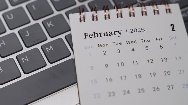 Close-up shot of a desk calendar on a laptop keyboard while a blue pen points to February 2026. Static camera highlights date indication and time planning in a clean office setting.