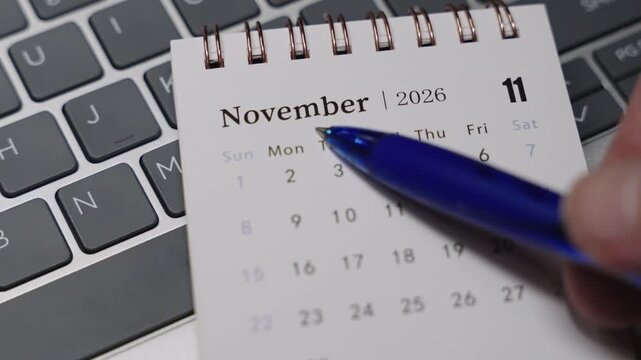 Close-up of a desk calendar on a laptop keyboard as a blue pen points to November 2026. Static camera presents clear date indication and monthly planning concept in a clean modern office environment.