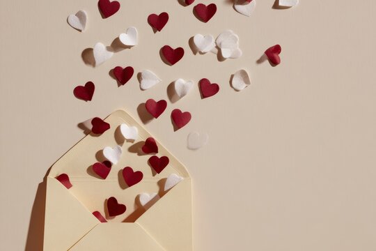 Numerous small red and white heart shapes emerge from the open flap of a pale colored envelope against a neutral background.