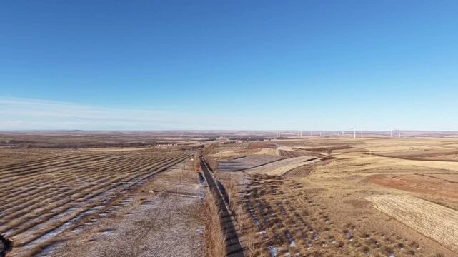 A cinematic aerial drone shot flying over a lonely country road cutting through vast harvested agricultural fields in winter. In the background, a row of wind turbines generates renewable energy under