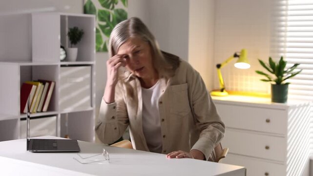 Amnesia. Senior woman with laptop trying to remember something at white table indoors