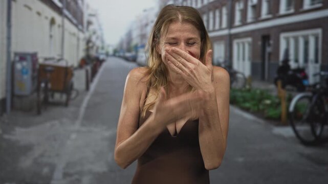 Woman with hand covering mouth and visible cleavage on street wearing brown dress; lighthearted amusement.