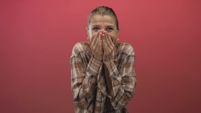 Woman smiling with hands covering mouth and wearing plaid shirt in studio with red backdrop; surprise warmth.