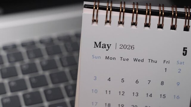 Close-up of a desk calendar on a laptop keyboard as a hand flips the page from April 2026 to May 2026. Camera stays static, showing a clear monthly time transition and structured planning concept ind