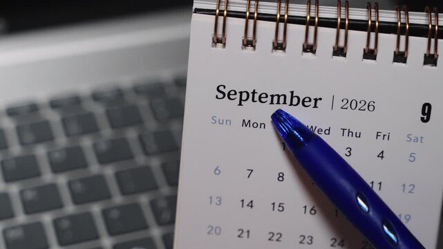 Close-up of a desk calendar placed on a laptop keyboard as a pen points to a specific date in September 2026. Camera remains static, presenting clear date selection and focused monthly planning conce