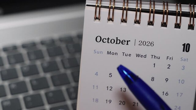 Close-up of a desk calendar placed on a laptop keyboard as a pen points to a specific date in October 2026. Camera remains static, presenting clear date selection and focused monthly planning concept