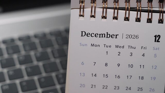 Close-up of a desk calendar on a laptop keyboard as a hand flips the page from November 2026 to December 2026. Camera stays static, presenting a clear monthly time transition and organized planning c