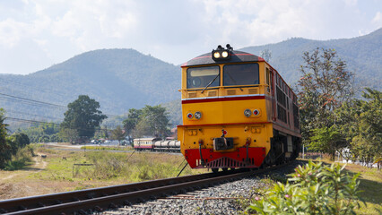 Obraz premium Yellow diesel locomotive train on railway track travels through scenic rural countryside. peaceful journey with beautiful mountain landscape in background