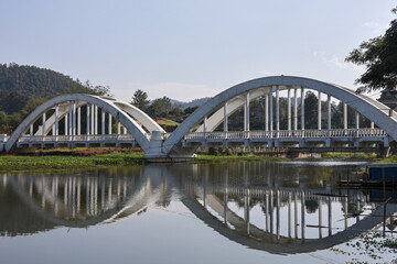 Obraz premium Scenic white railway bridge with two arches over tranquil river. peaceful structure and green mountain reflect perfectly in calm water on sunny day in Thailand