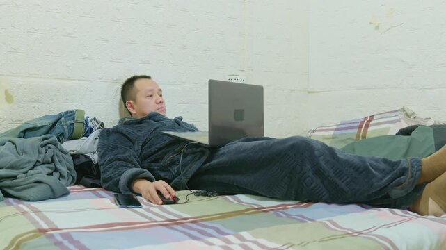 Asian man wearing pajamas playing on his laptop in bed. Asian man playing computer games in bed. Man stay up late playing computer games instead of sleeping at night.