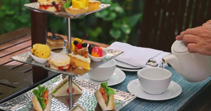 Person pouring hot tea from white teapot into ceramic cup beside three-tier silver stand filled with gourmet sandwiches, pastries and cakes in outdoor garden setting, elegant atmosphere, luxury hospit