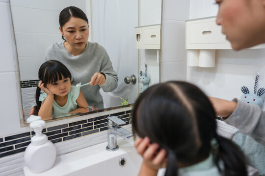 stubborn child girl refusing to brush her teeth in the bathroom. mother is trying to encourage her unhappy daughter during the morning routine, while child looks grumpy and resistant.