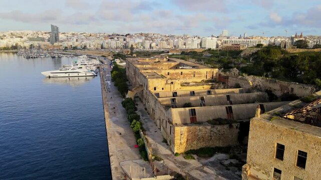 Drone view of abandoned Lazzaretto buildings along Marsamxett Harbour with marina and city skyline in Malta.