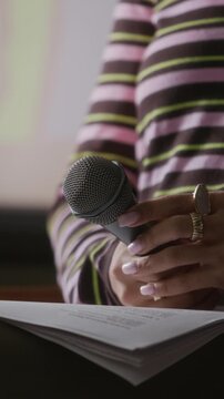 Vertical cropped view of unrecognizable speaker seated on stage holding notes and microphone delivering talk to audience at public event