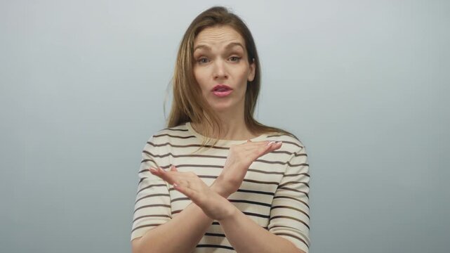 Woman in striped shirt with serious expression crossing arms x gesture in light blue studio; rejection.