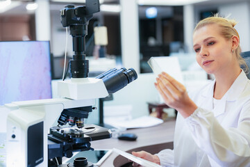 Serious Caucasian female scientist inspecting glass slide near microscope in modern lab. Young...