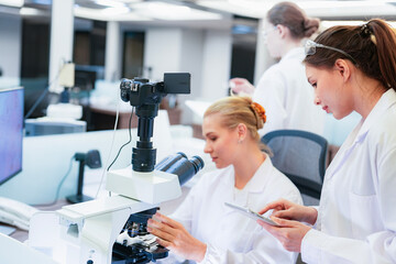 Asian female scientist using tablet to discuss histology results with colleague at microscope. Team...