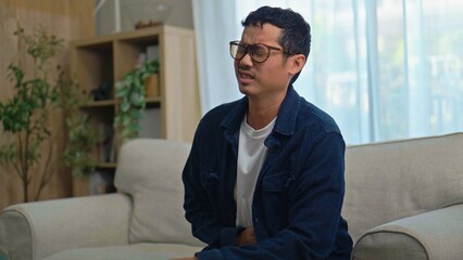 Fototapeta premium Asian Man Suffering from Stomach Pain and Abdominal Cramps on Sofa at Home.