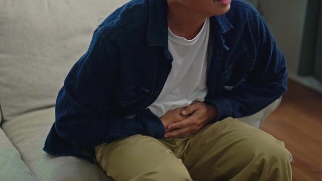 Asian Man Suffering from Stomach Pain and Abdominal Cramps on Sofa at Home.