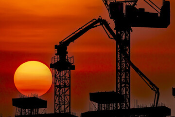 Silhouetted construction cranes and building structures against vibrant orange sunset create dramatic urban scene. sun sets behind cranes, casting warm glow