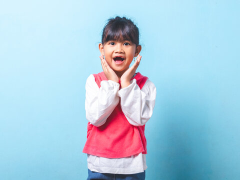 A cute toothless little girl standing facing forward holding cheeks with amazing expression on isolated blue background for dental promotion or surprise sale announcement.