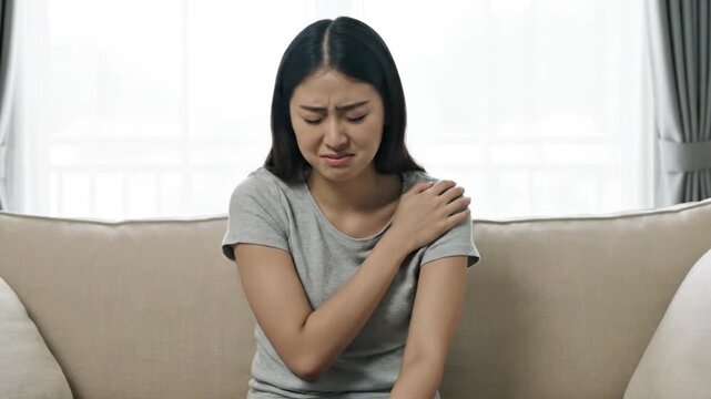 Asian Woman Shoulder Pain on Couch &ndash; Female Holding Painful Shoulder Discomfort at Home